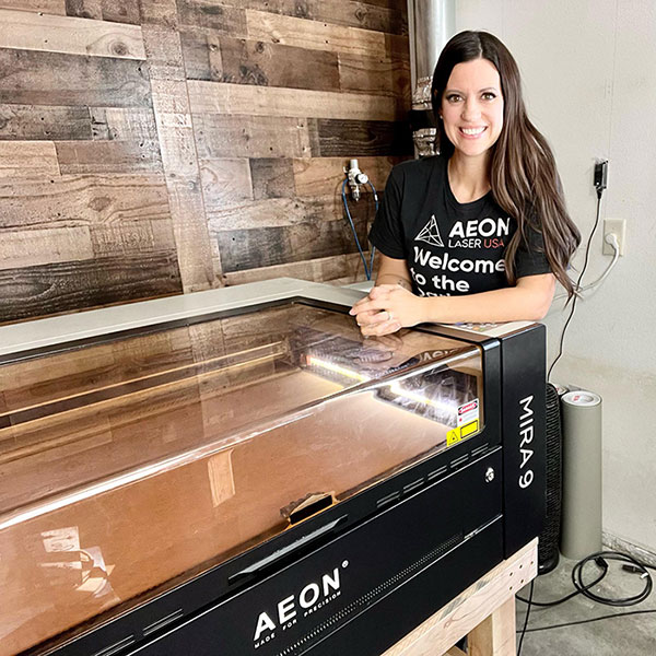 women in her craft room, posing by her Aeon CO2 laser. Her t-shirt reads, welcome to the dark side!