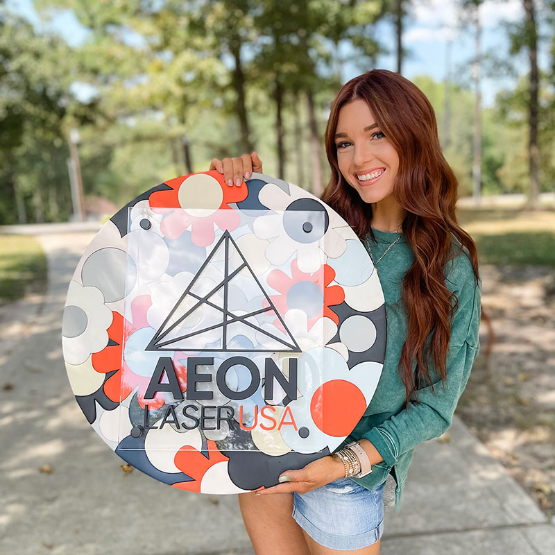 photo of a women with wavy light brown hair, smiling and holding a custom made round wooden business sign, in a natural outdoor setting