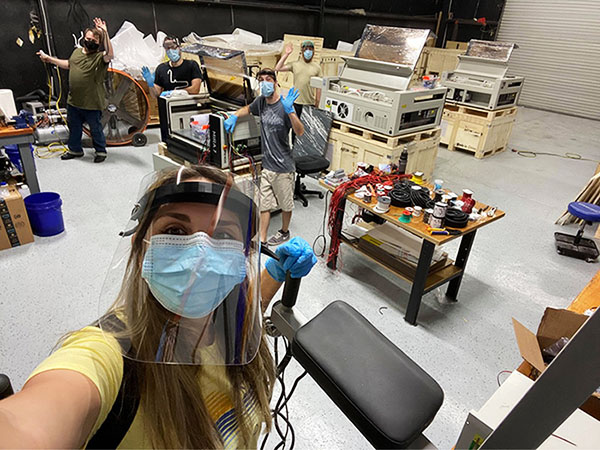 a woman taking a selfie at the Aeon Laser USA warehouse, workers waving with machines in the background