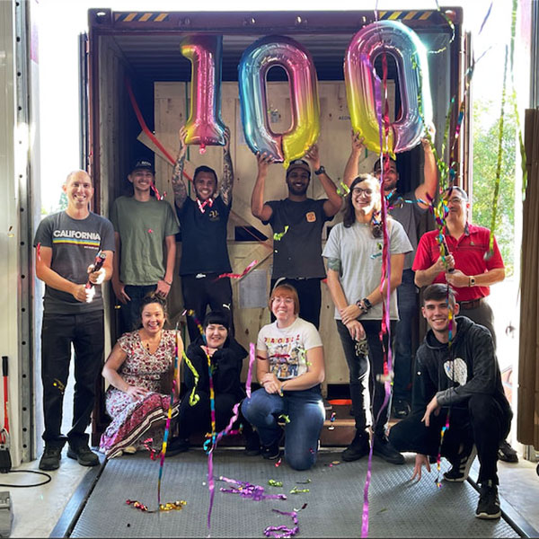 group of employees gathered in front of a loading dock filled with machines from the 100th shipment, with balloons overhead celebrating the milestone.
